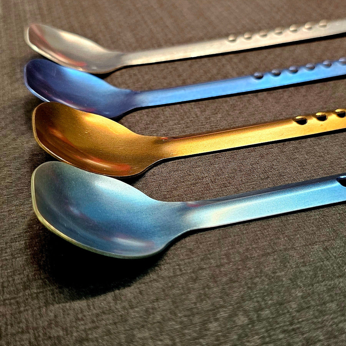 Four metal spoons in different colors arranged on a textured surface.