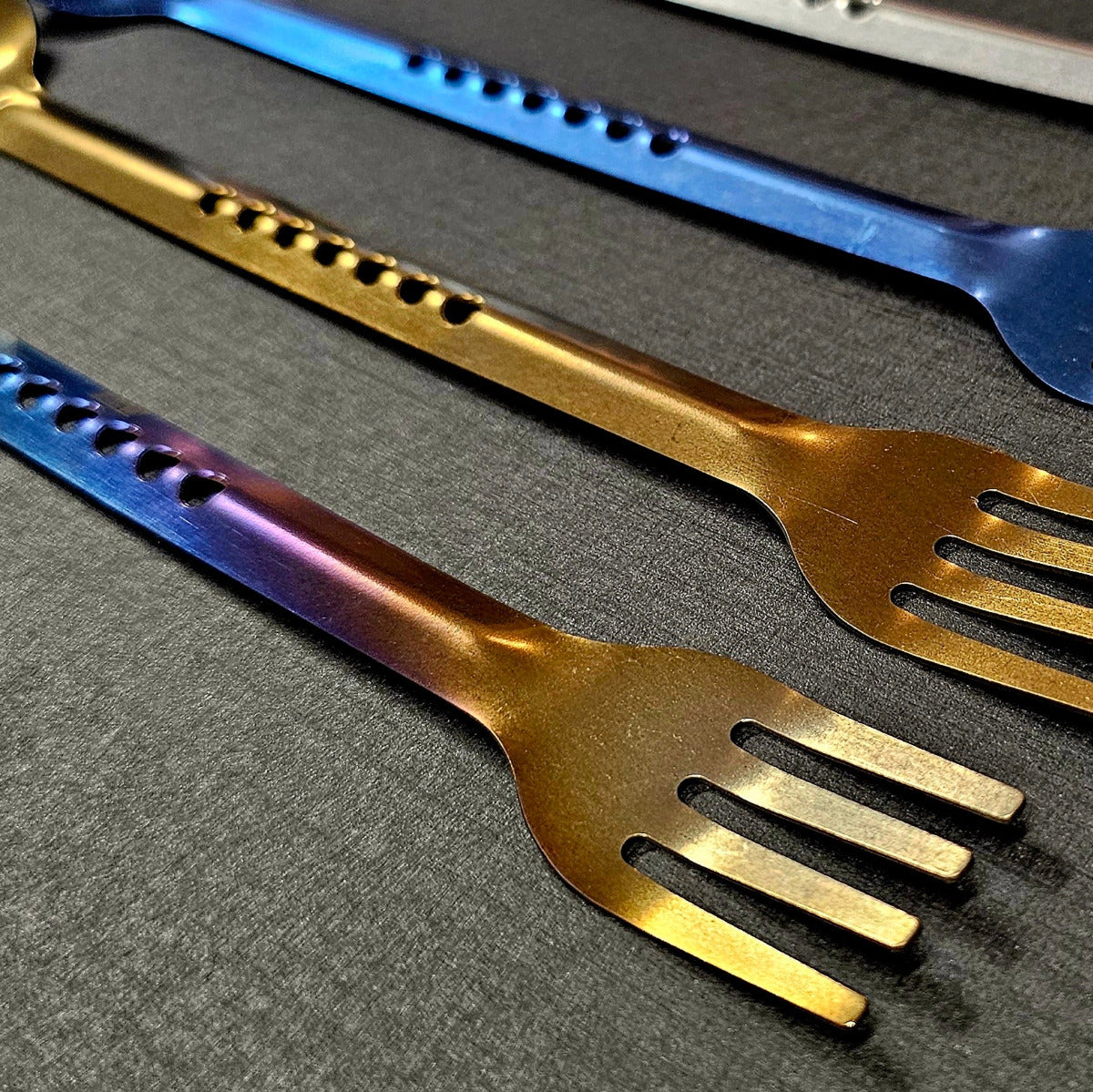 Set of colorful metal utensils including forks and a spoon on a dark surface.